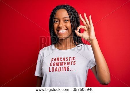 Young african american woman wearing sarcasm coments text on t-shirt over red background doing ok sign with fingers, excellent symbol