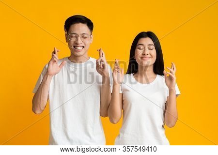 Making Cherished Wish. Young Asian Couple With Closed Eyes And Crossed Fingers Over Yellow Backgroun