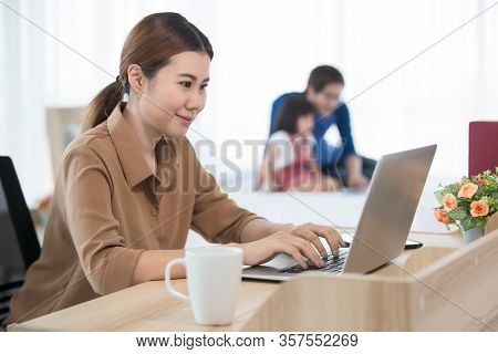 Asian Businesswoman Mother Working At Home With Notebook Computer, Tablet, And Smartphone While Her 