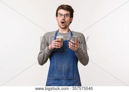 Portrait Of Shocked Guy In Stupor, Wearing Apron As Working In Coffee Shop Or Bar Counter, Hold Mobi