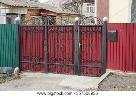Red Metal Gate With Black Wrought Iron Rods And An Iron Fence On The Street On A Sunny Day