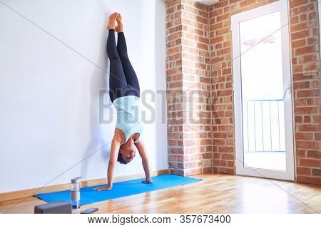 Middle age beautiful sportwoman smiling happy on mat. Practicing yoga doing wall-assisted handstand pose at gym