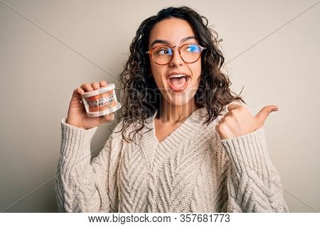 Young beautiful woman with curly hair holding plastic teeth with dental baces pointing and showing with thumb up to the side with happy face smiling