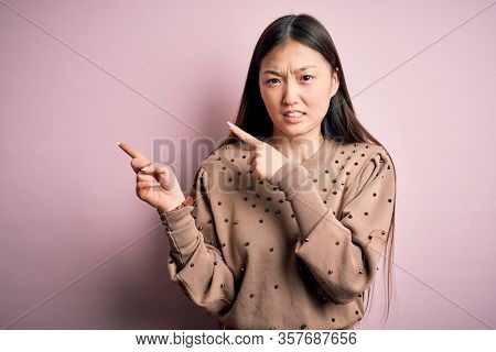 Young beautiful asian woman wearing fashion and elegant sweater over pink solated background Pointing aside worried and nervous with both hands, concerned and surprised expression