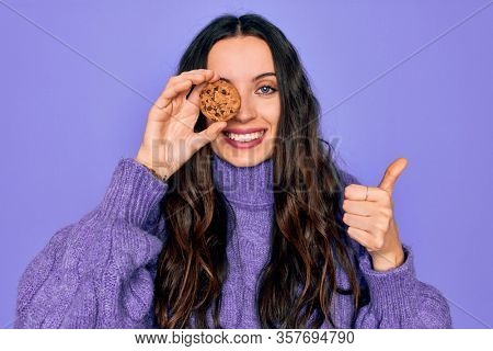 Young beautiful woman with blue eyes holding chocolate sweet coockie over eye happy with big smile doing ok sign, thumb up with fingers, excellent sign