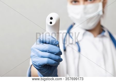 Close-up shot of doctor wearing protective surgical mask ready to use infrared forehead thermometer (thermometer gun) to check body temperature for virus symptoms - epidemic virus outbreak concept
