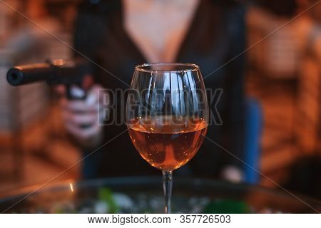 Glass Of Alcohol Close Up. Behind Him The Silhouette Of A Woman With A Gun.