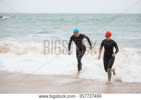 Swimmers In Wetsuits Running In Sea Waves. High Angle View Of Athletic People In Sportswear Running 