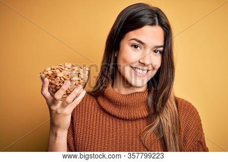 Young beautiful girl holding bowl of salty peanuts standing over yellow background with a happy face standing and smiling with a confident smile showing teeth