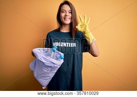 Beautiful volunteer girl caring environment doing volunteering holding bag with rubish bottles doing ok sign with fingers, excellent symbol