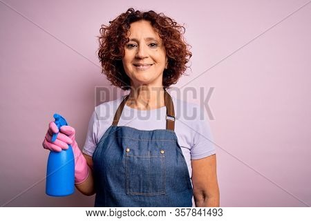 Middle age curly hair woman cleaning doing housework wearing apron and gloves using spayer with a happy face standing and smiling with a confident smile showing teeth