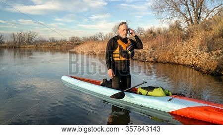 A senior male paddler wearing wetsuit and life jacket is making a phone call after stand up paddling workout on a calm lake in Colorado.
