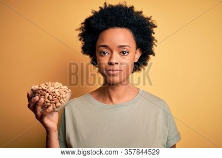 Young African American afro woman with curly hair holding bowl with salty peanuts with a confident expression on smart face thinking serious
