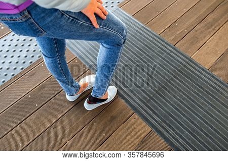 Female Legs In Denim Jeans Pants And Light Summer Sneakers Standing On Wooden Floow.