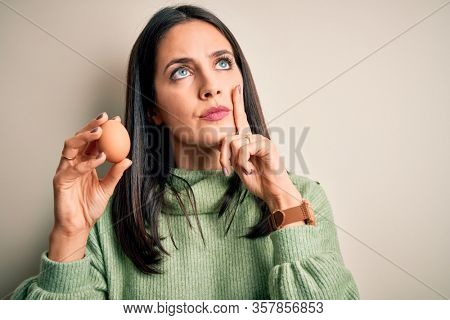 Young brunette woman with blue eyes holding fresh raw egg over isolated background serious face thinking about question, very confused idea