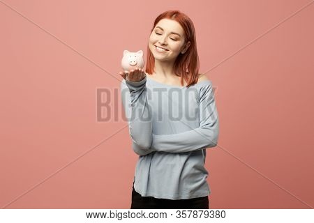 Smiling ginger girl with piggy bank in hands standing in studio isolated