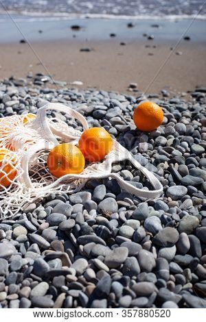 White Cotton Eco String Bag With Oranges On A Rocky Beach. Sea View With A Blue Sky. Zero And Eco Wa
