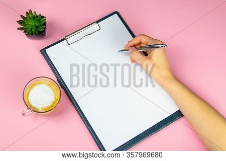 Tablet With White Flattery Next To Which Stands A Green Plant, Hand Holds A Pen And Writes On A Shee