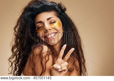 Cheerful Pretty Latin Female With Gold Hydrogel Patches Under Closed Eyes Show V-sigh Gesture Posing