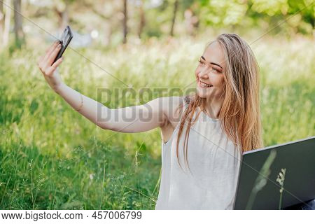 Girl Sits Outdoors And Works At A Laptop. Makes A Selfie On The Phone. Freelance. Selfeducation. The