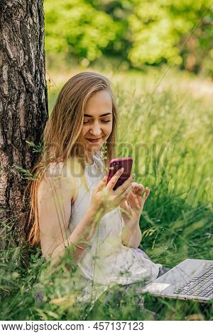 Girl Sitting On The Grass Looking At The Phone. Freelance. Selfeducation. The Concept Of Remote Lear