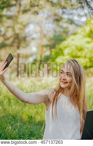 Girl Sits Outdoors And Works At A Laptop. Makes A Selfie On The Phone. Freelance. Selfeducation. The