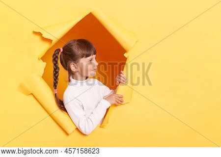 Side View Of Caucasian Positive Little Girl With Braids Wearing White Turtleneck Posing In Torn Hole