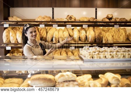 Portrait Of Female Bakery Worker Showing Fresh Baked Pastries And Bread On Shelf In Bakery Shop.