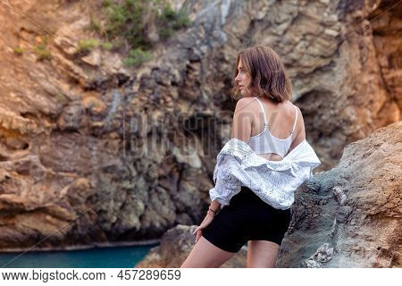 A Beautiful European-looking Girl In A White Shirt And Black Shorts Poses Against The Backdrop Of Ro