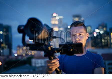 Man Filming With Camera And Gimbal. Portrait Of Videographer Against Illuminated Urban Skyline At Ni