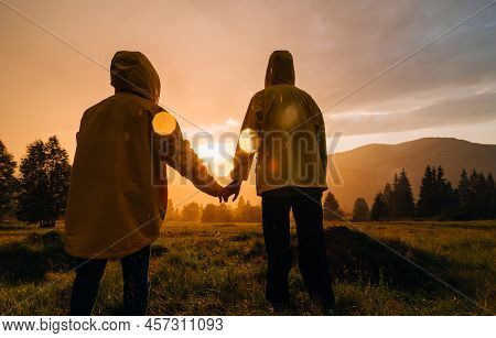 Couple In Yellow Raincoats Stand Holding Hands On A Meadow In The Mountains On A Background Of Orang