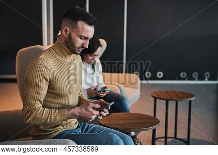 Concentrated Multitasking Young Caucasian Man Using Mobile Phone, Booking A Hotel, Sitting Next To H