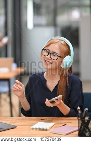 Happy Woman Employee Wearing Headphones Enjoy Favorite Song And Using Smart Pone At Office Desk
