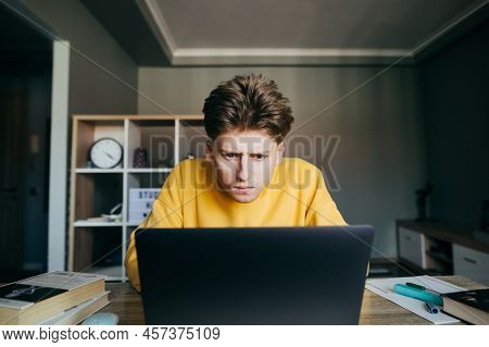 Pensive Young Male Student Sitting At Home With Books And Notebooks Studying Remotely Online, Lookin