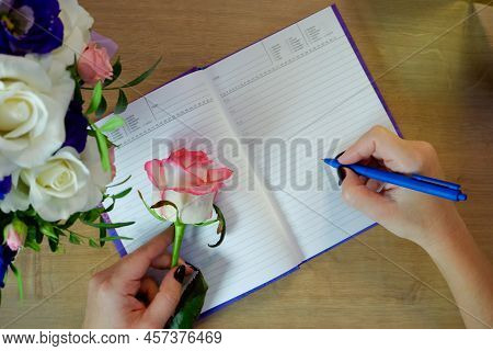 Female Hands Writing In Open Notebook And Bouquet Of Roses On Old Wooden Table. Planning Notebook Wi