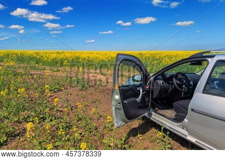 Small Silver Car In Rapseed Field With Open Door At Sunny Noon.