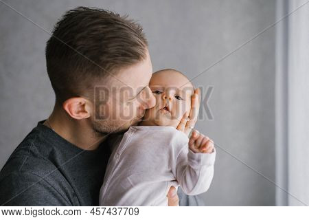 Dad Holds His Baby In His Arms At Home And Kisses The Baby On The Cheek