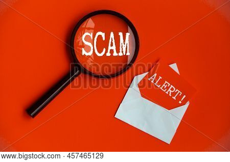 Scam Alert Text On Magnifying Glass And Notepad On Red Cover Background. Scamming And Fraud Concept