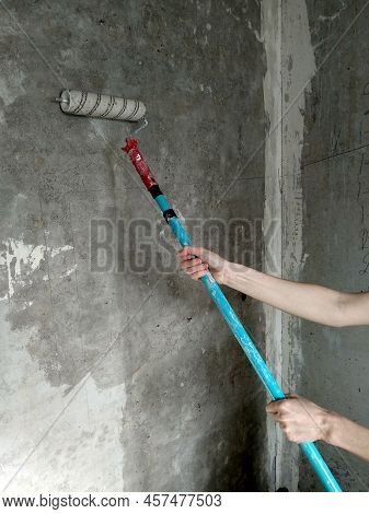 Woman Hands With A Long-handled Roller Applying A Primer To The Gray Empty Wall