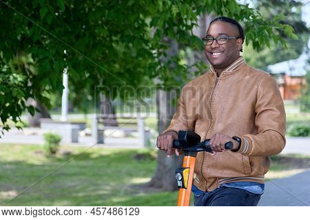 Close-up Of A Dark Skinned Smiling Man With Glasses And A Brown Jacket Driving An Electric Scooter. 