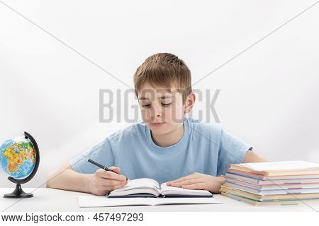 Boy Of 8-9 Years Old Does Homework. Schoolboy Sits At Table With Book, Notebooks And Globe. Isolated