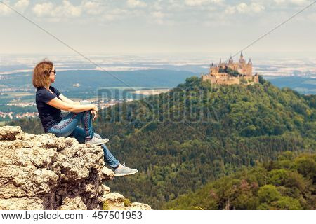 Girl Tourist Sitting On Cliff Looks At Hohenzollern Castle On Mountain Top, Germany. Woman Travels I