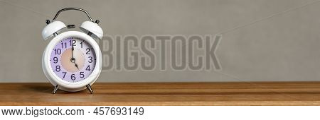 Five Oclock On The Alarm. A White Alarm Clock Stands On A Wooden Table. The Clock Hand Points To Fiv