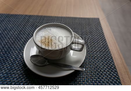 A White Cup Hot Capuccino With White Bubble Mug, Stanless Spoon On Ceramic Plate On Brown Wooden Tab