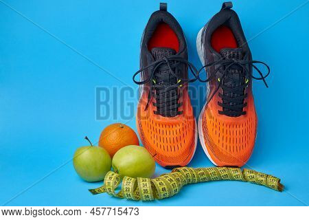 Orange Sneakers, Measuring Tape, Water Botlle And Fruits Apples And Oranges On A Blue Background