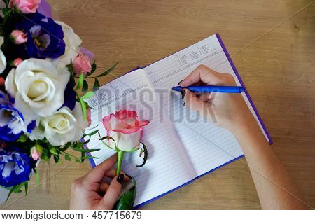 Female Hands Writing In Open Notebook And Bouquet Of Roses On Old Wooden Table. Planning Notebook Wi