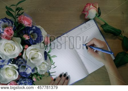 Female Hands Writing In Open Notebook And Bouquet Of Roses On Old Wooden Table. Planning Notebook Wi