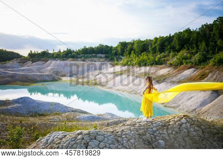 A Small 6-7 Years Old Ukrainian Girl In A Yellow Dress With Wings In Yellow Fabric By The Lake. Blue