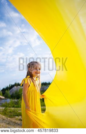 A Small 6-7 Years Old Ukrainian Girl In A Yellow Dress With Wings In Yellow Fabric By The Lake. Blue