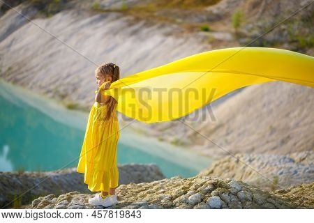 A Small 6-7 Years Old Ukrainian Girl In A Yellow Dress With Wings In Yellow Fabric By The Lake. Blue